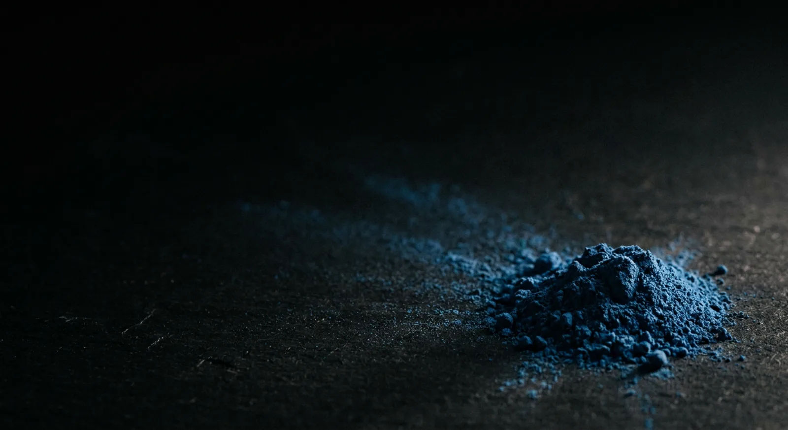 Gundukan pigmen bubuk biru azure di atas permukaan hitam matte, disinari cahaya menyamping, dengan serakan butiran halus di sekitarnya.
